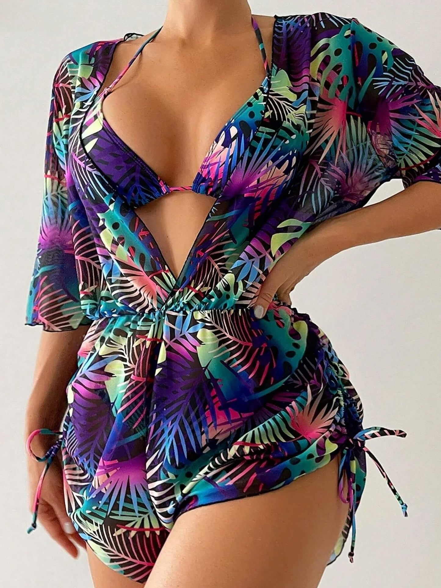 Purple - A woman models a sheer, short-sleeve mini cover-up with a vibrant tropical leaf print in shades of purple, blue, and pink, featuring a deep V-neck, drawstring waist, and ruched sides with ties.