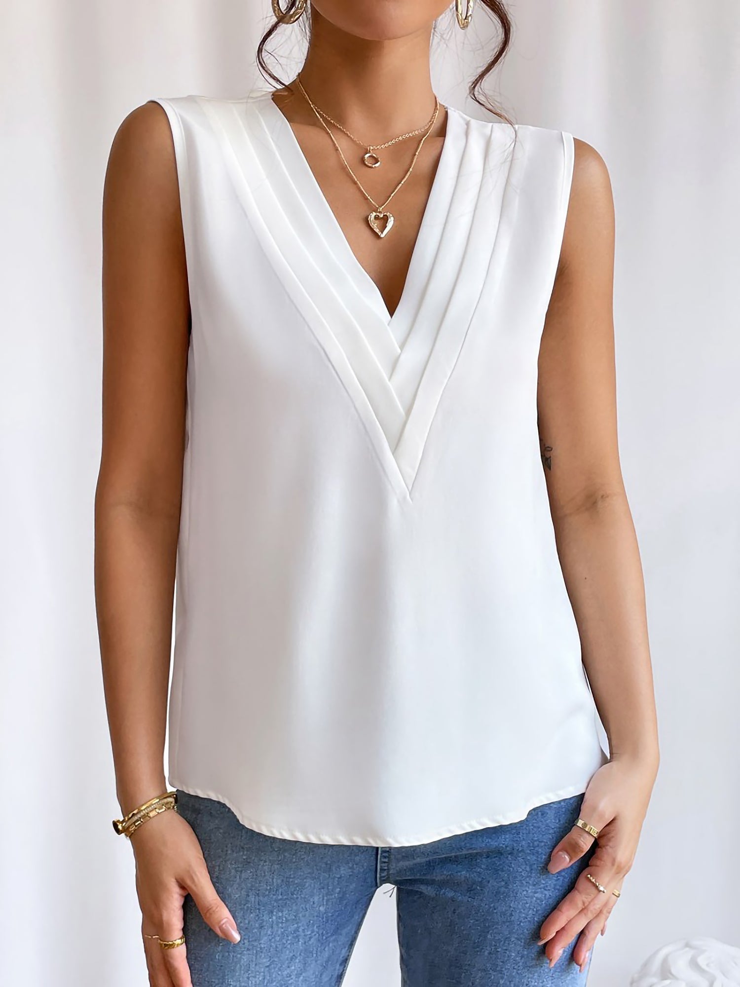 White - Women's solid white sleeveless blouse featuring a deep V-neckline with layered pleat detailing, styled with blue jeans and a black belt.