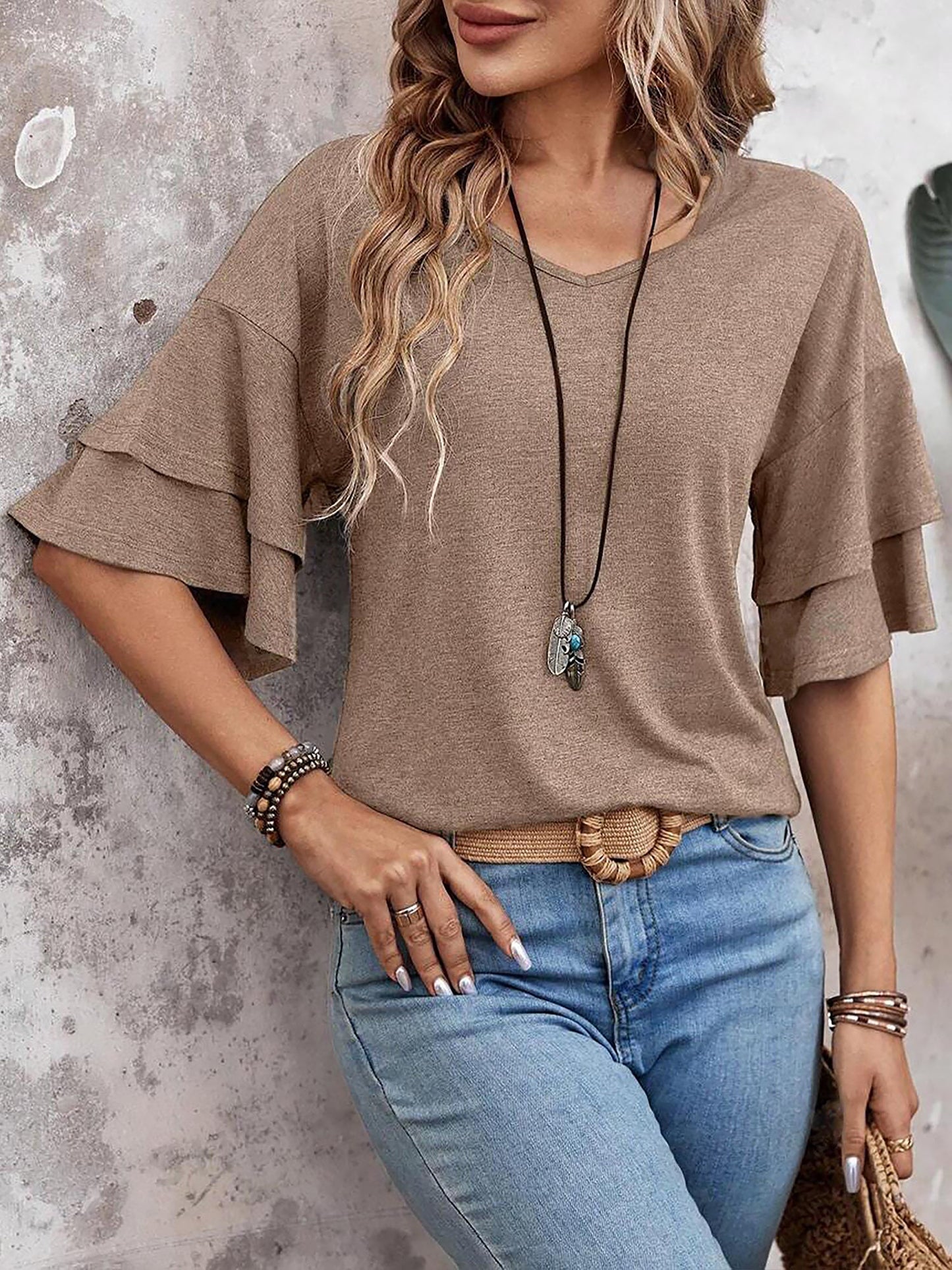 Camel - Women's solid pink V-neck blouse featuring tiered ruffle short sleeves, styled with blue jeans and a long pendant necklace.