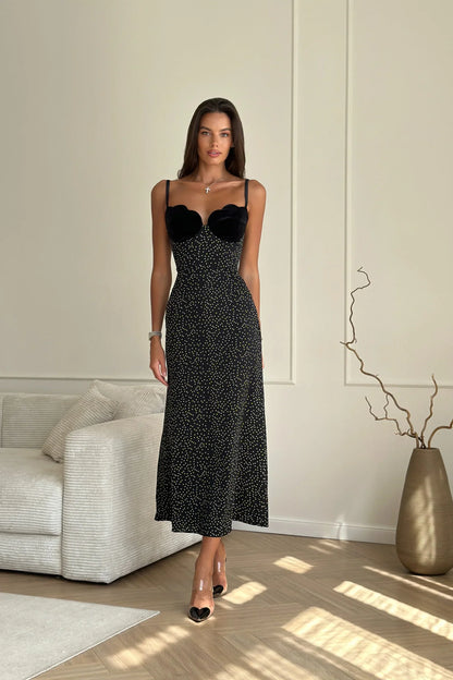 Black - Woman wearing an ankle-length polka dot dress featuring a velvet sweetheart bodice and delicate spaghetti straps, leaning against a white wall in a brightly lit minimalist room.