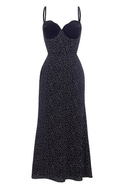 Black - Woman wearing an ankle-length polka dot dress featuring a velvet sweetheart bodice and delicate spaghetti straps, leaning against a white wall in a brightly lit minimalist room.