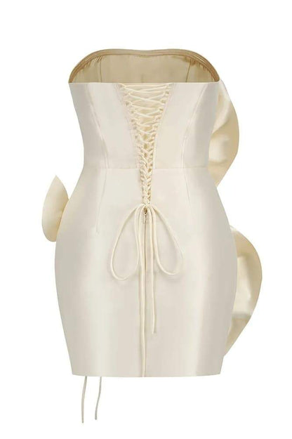 A woman models a strapless, ivory satin mini dress featuring two large, sculptural rosette appliques down the front against a neutral background.
