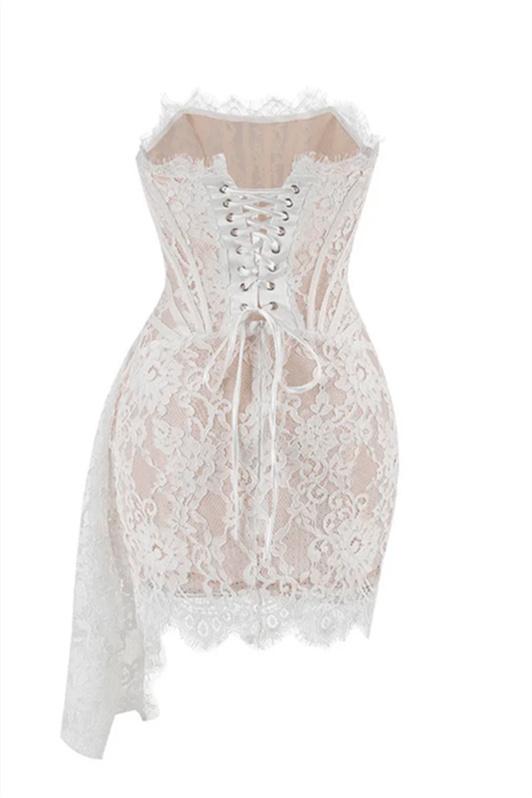 A woman models a white strapless lace mini dress while standing on a grand marble staircase. The dress features a sheer corset bodice with visible boning and a draped, asymmetrical skirt.