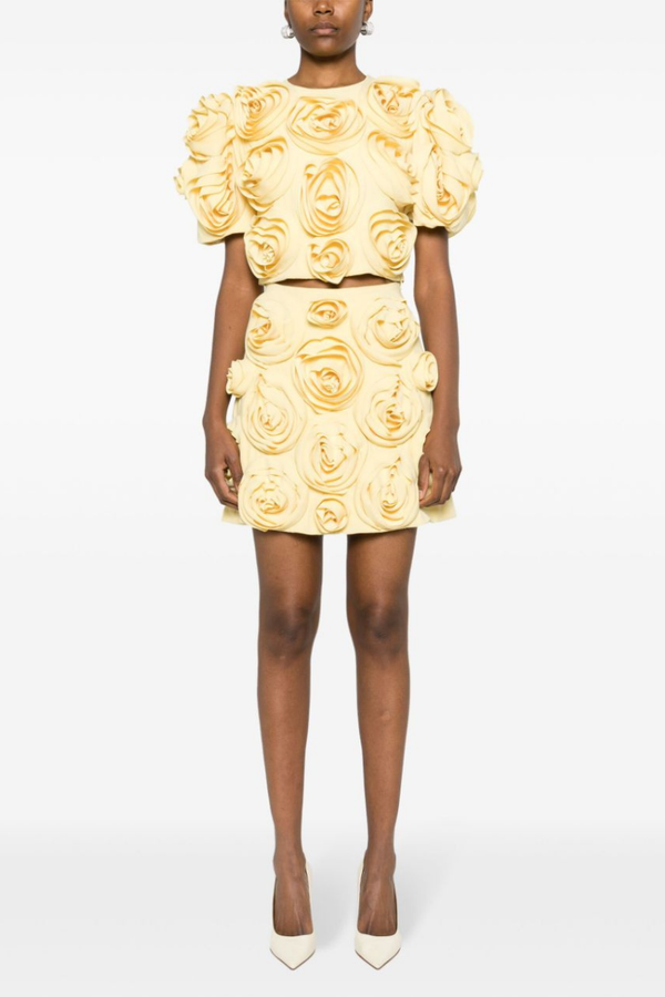 A woman wearing a pale yellow two-piece set featuring a cropped top with short puff sleeves and a matching mini skirt, completely covered in large 3D fabric rosette appliqués.