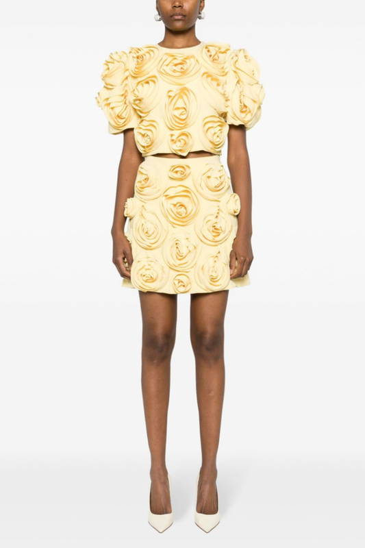 A woman wearing a pale yellow two-piece set featuring a cropped top with short puff sleeves and a matching mini skirt, completely covered in large 3D fabric rosette appliqués.