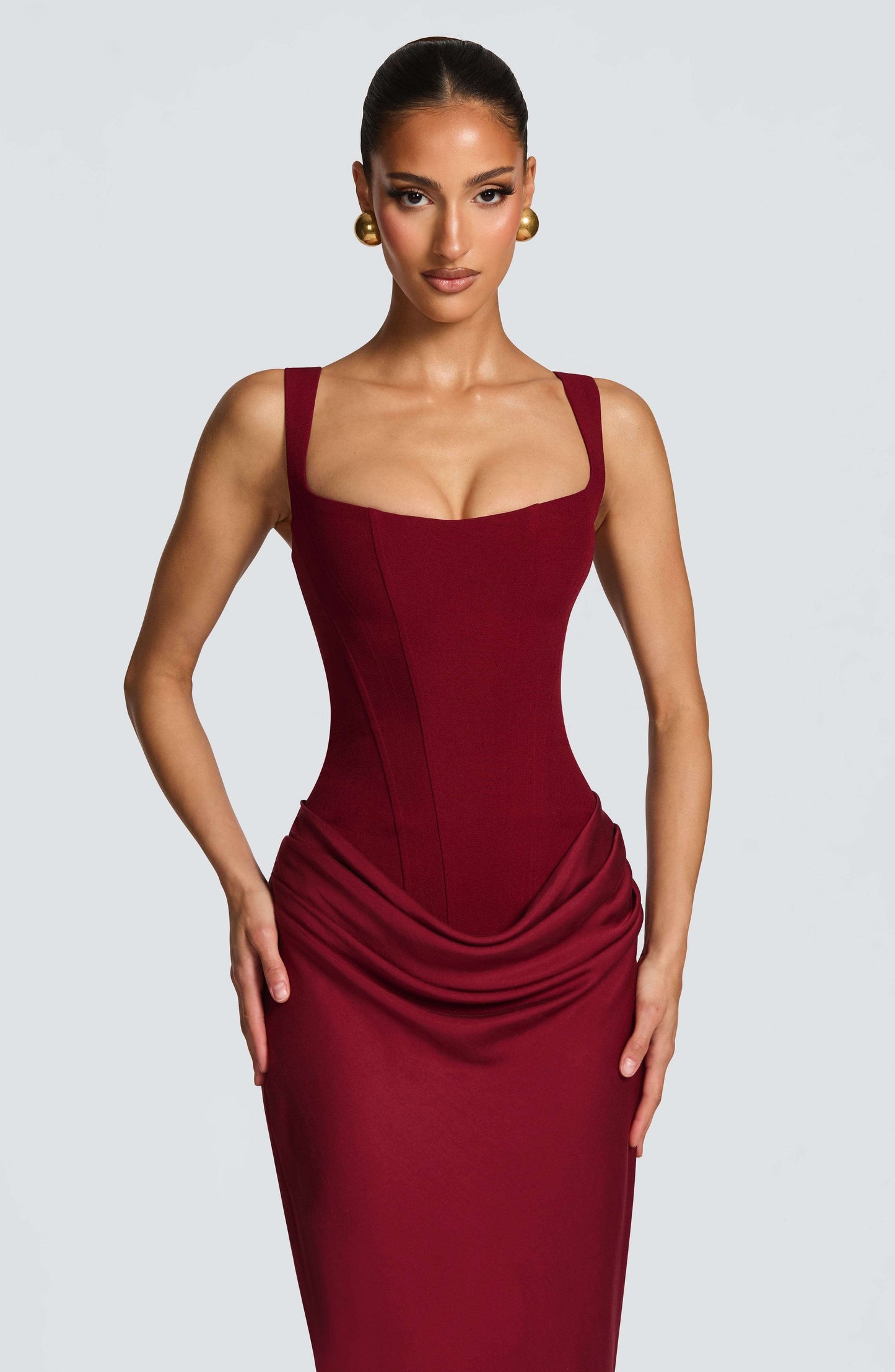 Black - A woman stands in an elegant, dimly lit setting wearing a deep red, floor-length formal dress featuring a structured corset bodice, wide straps, and elegant draping at the hips.