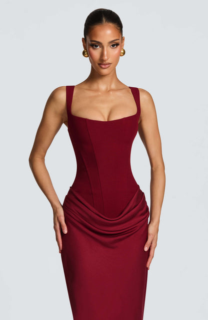 Black - A woman stands in an elegant, dimly lit setting wearing a deep red, floor-length formal dress featuring a structured corset bodice, wide straps, and elegant draping at the hips.