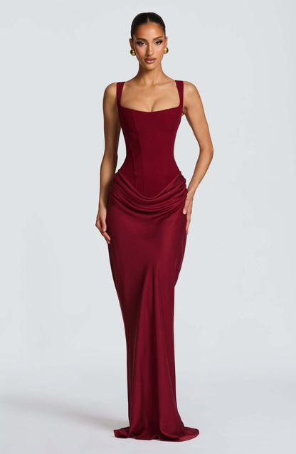 Black - A woman stands in an elegant, dimly lit setting wearing a deep red, floor-length formal dress featuring a structured corset bodice, wide straps, and elegant draping at the hips.