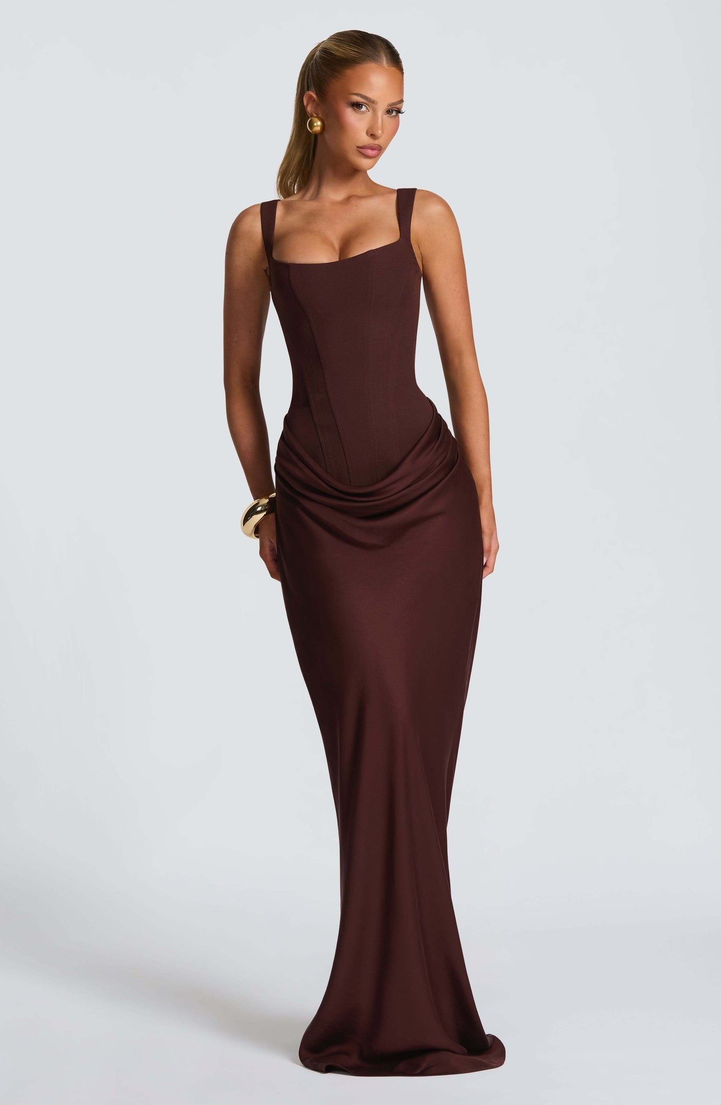 Black - A woman stands in an elegant, dimly lit setting wearing a deep red, floor-length formal dress featuring a structured corset bodice, wide straps, and elegant draping at the hips.