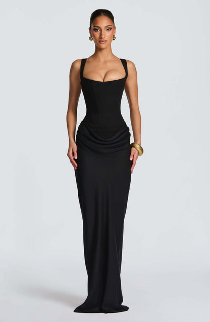 Black - A woman stands in an elegant, dimly lit setting wearing a deep red, floor-length formal dress featuring a structured corset bodice, wide straps, and elegant draping at the hips.