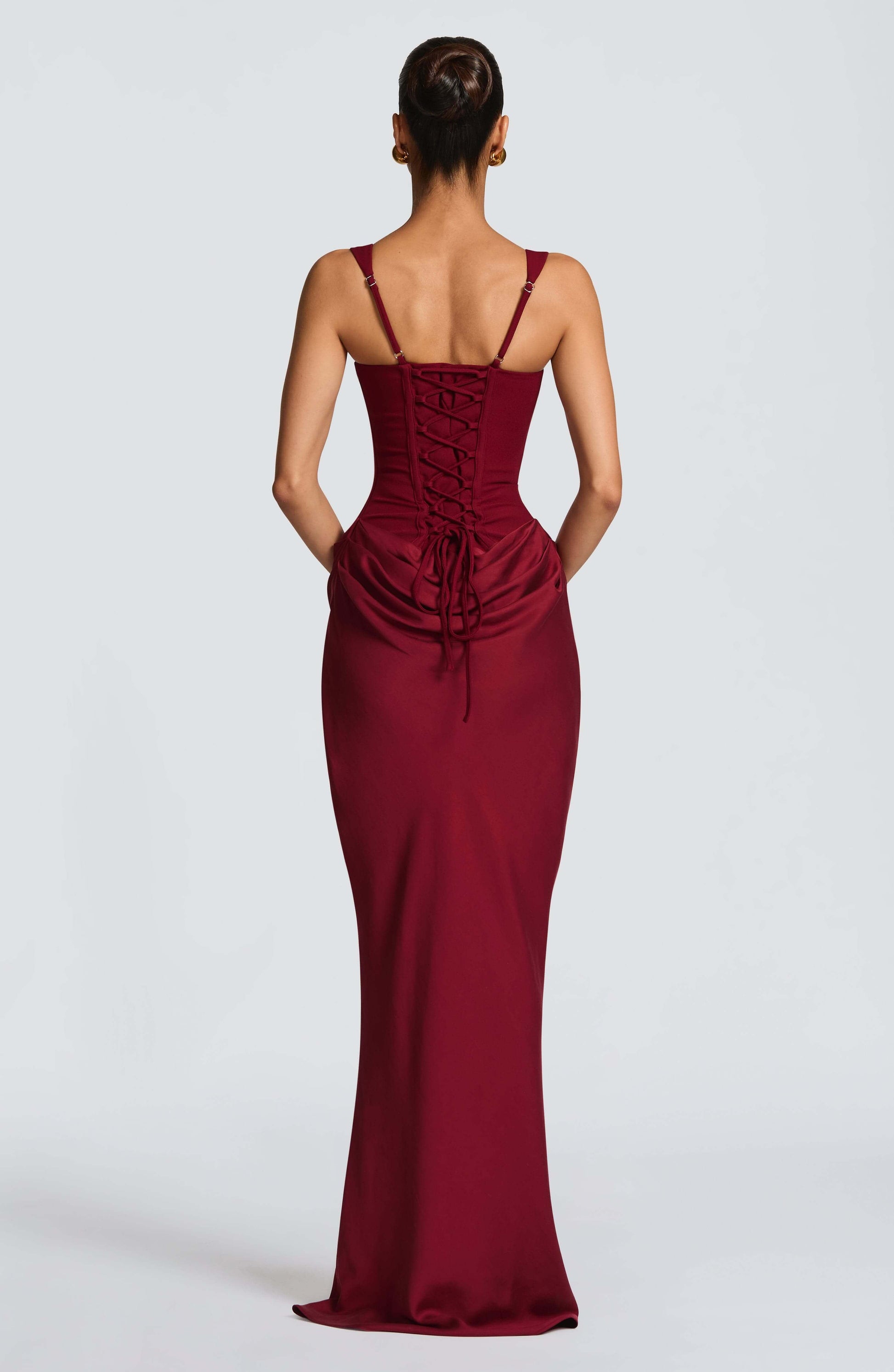 Black - A woman stands in an elegant, dimly lit setting wearing a deep red, floor-length formal dress featuring a structured corset bodice, wide straps, and elegant draping at the hips.