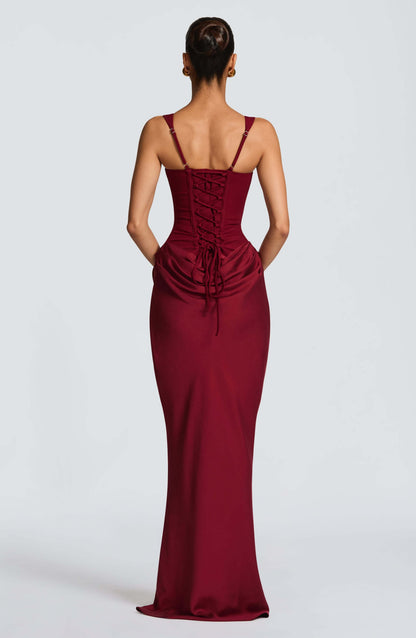 Black - A woman stands in an elegant, dimly lit setting wearing a deep red, floor-length formal dress featuring a structured corset bodice, wide straps, and elegant draping at the hips.