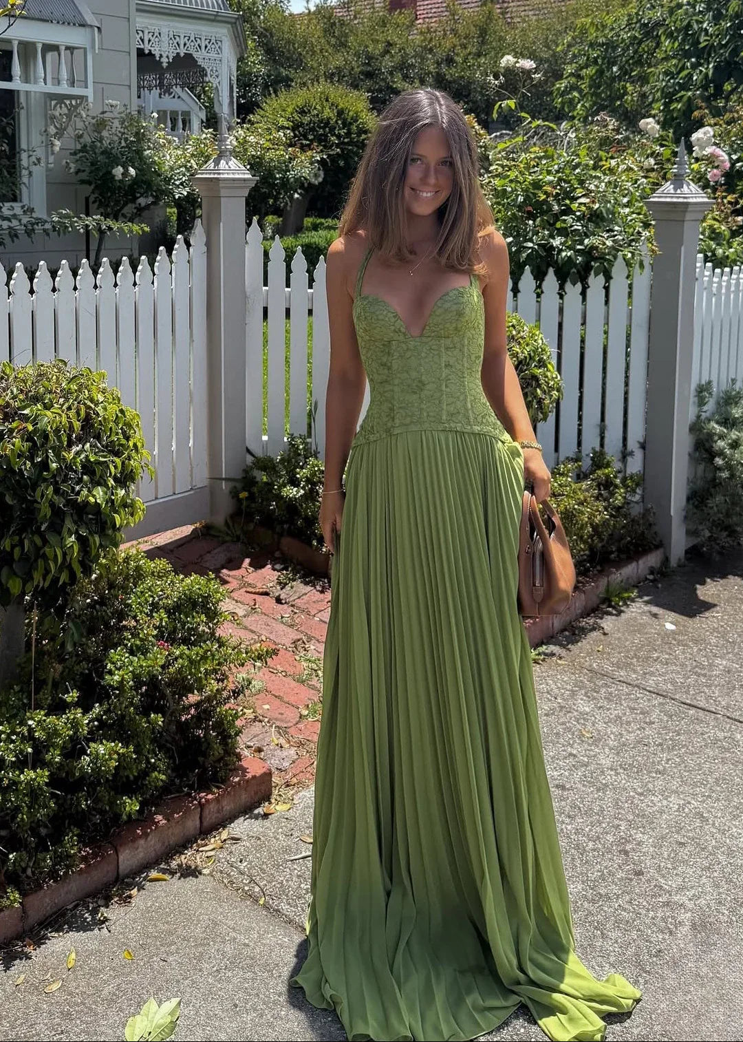 Green - A woman wearing an elegant olive green maxi dress featuring a structured lace corset bodice, halter neckline, and a sweeping floor-length pleated skirt, standing outdoors near stone steps and tropical foliage.