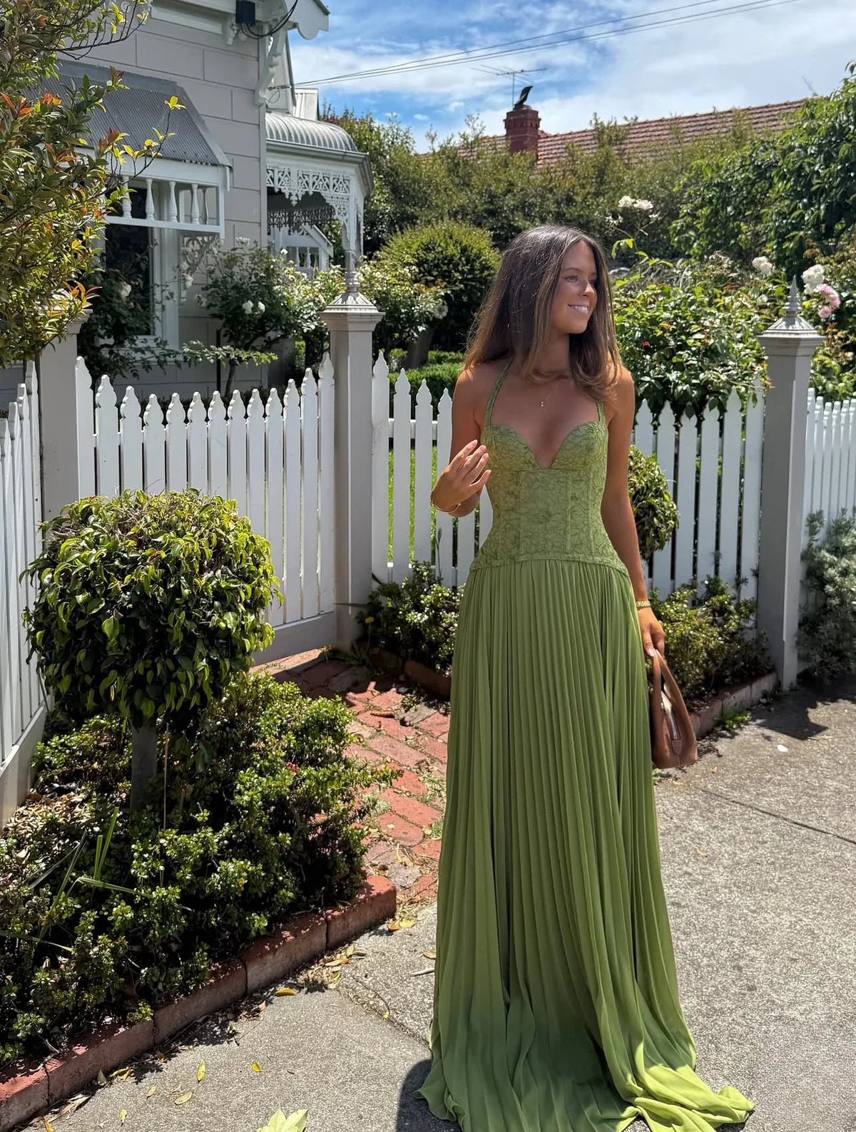 Green - A woman wearing an elegant olive green maxi dress featuring a structured lace corset bodice, halter neckline, and a sweeping floor-length pleated skirt, standing outdoors near stone steps and tropical foliage.