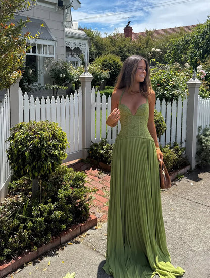 Green - A woman wearing an elegant olive green maxi dress featuring a structured lace corset bodice, halter neckline, and a sweeping floor-length pleated skirt, standing outdoors near stone steps and tropical foliage.