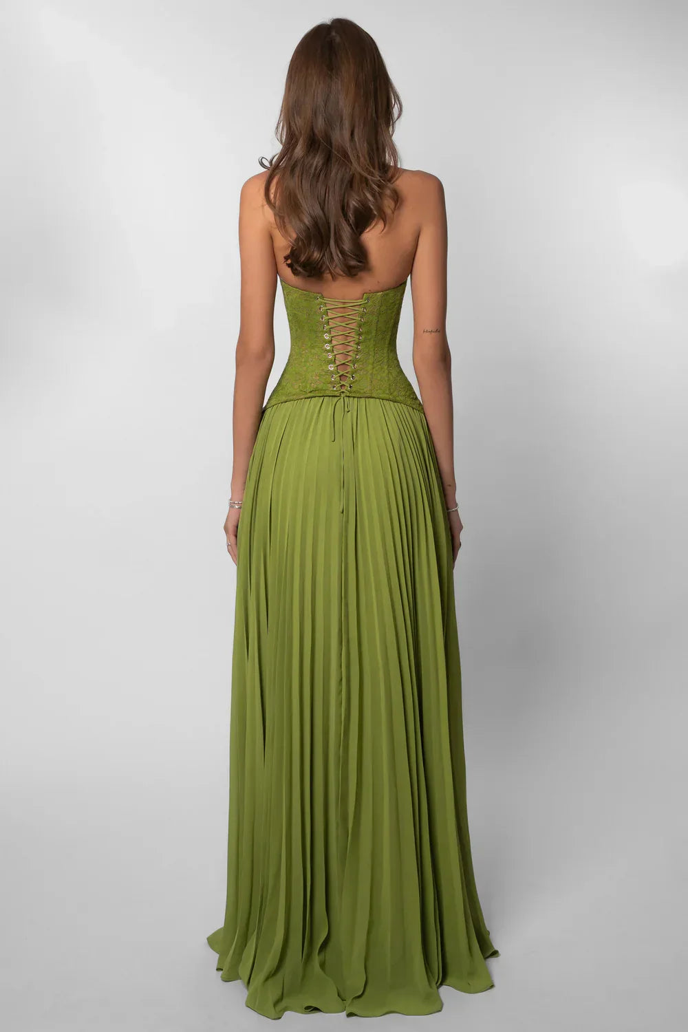 Green - A woman wearing an elegant olive green maxi dress featuring a structured lace corset bodice, halter neckline, and a sweeping floor-length pleated skirt, standing outdoors near stone steps and tropical foliage.