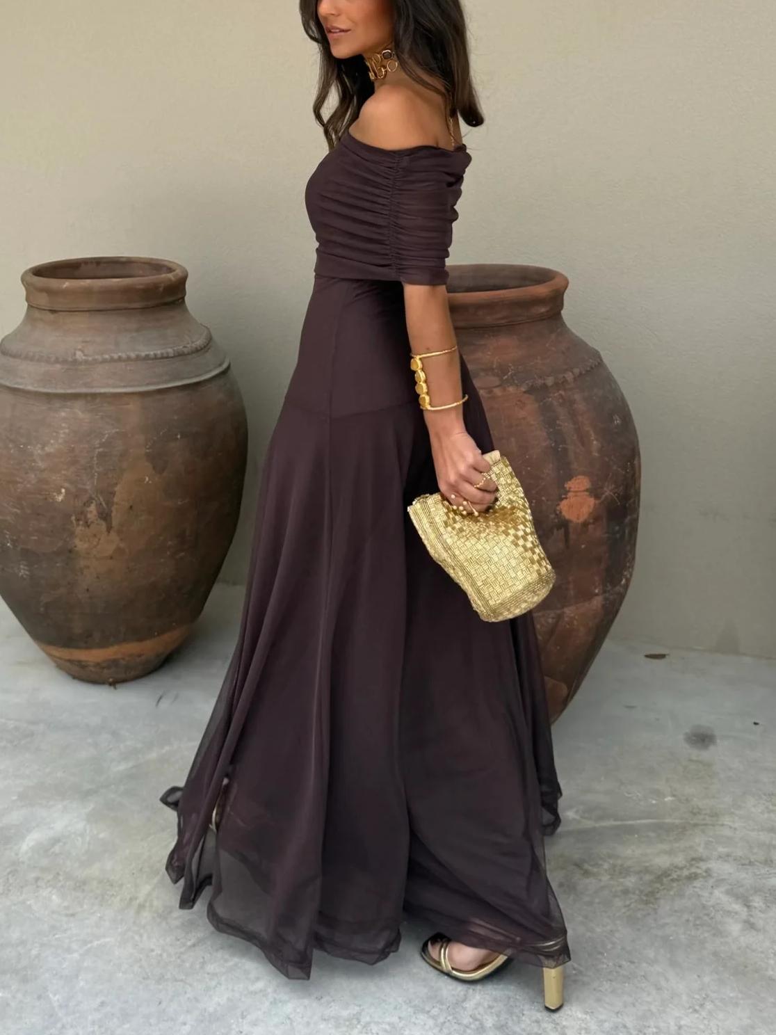 Brown - A woman wearing a dark brown, formal off-the-shoulder maxi dress featuring a ruched neckline and a flowing sheer mesh overlay over a fitted underlay, styled with gold accessories.