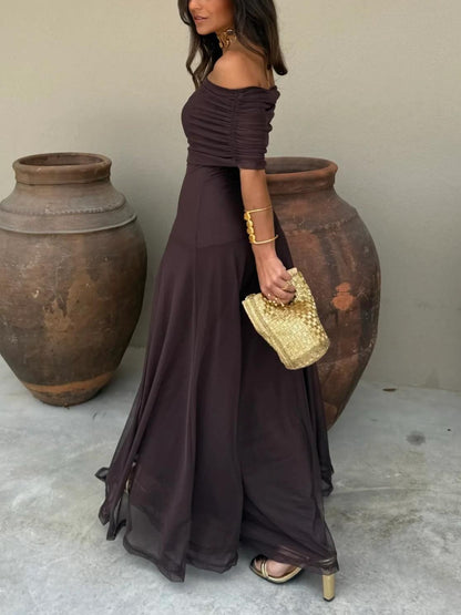 Brown - A woman wearing a dark brown, formal off-the-shoulder maxi dress featuring a ruched neckline and a flowing sheer mesh overlay over a fitted underlay, styled with gold accessories.