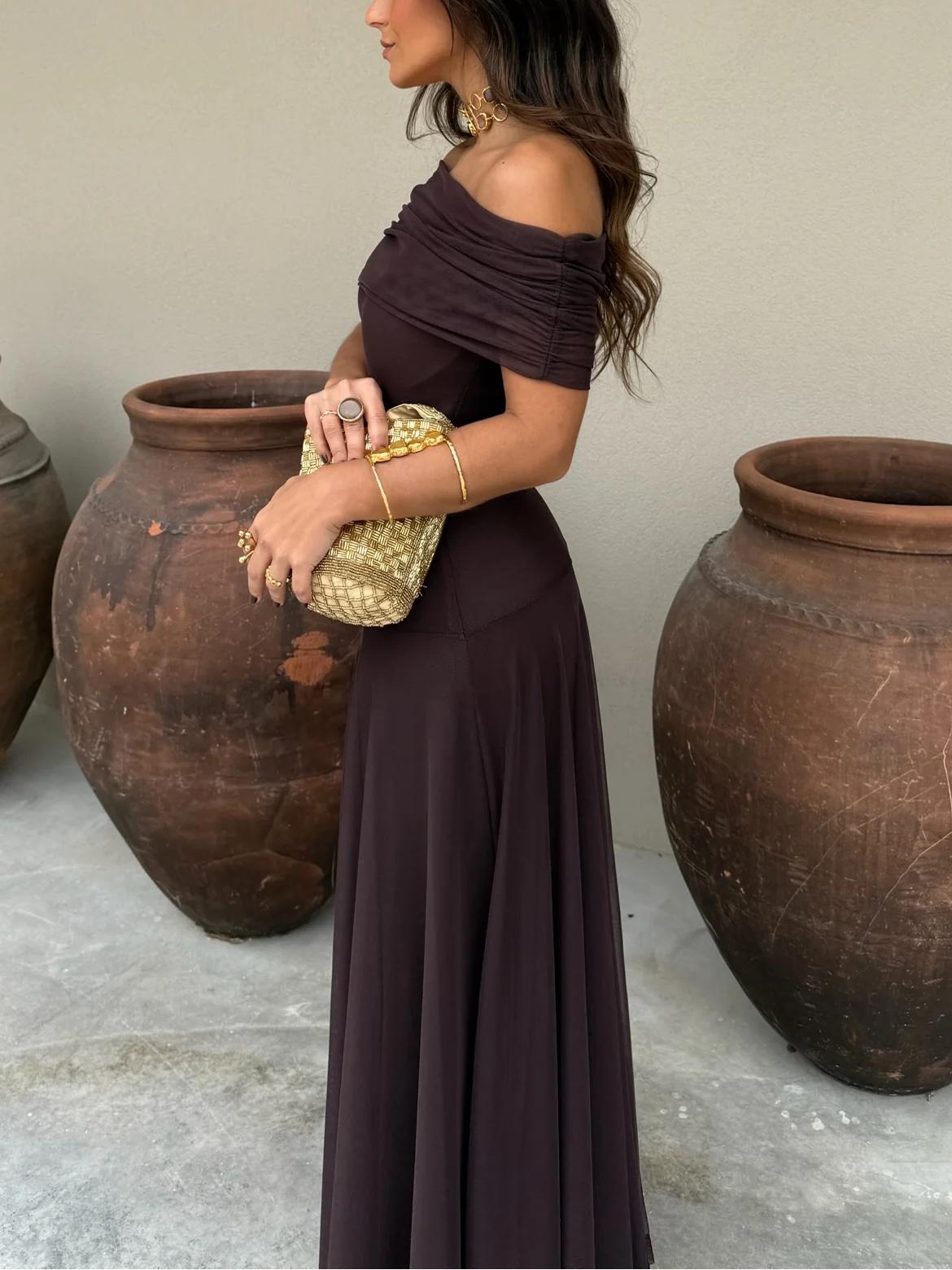 Brown - A woman wearing a dark brown, formal off-the-shoulder maxi dress featuring a ruched neckline and a flowing sheer mesh overlay over a fitted underlay, styled with gold accessories.