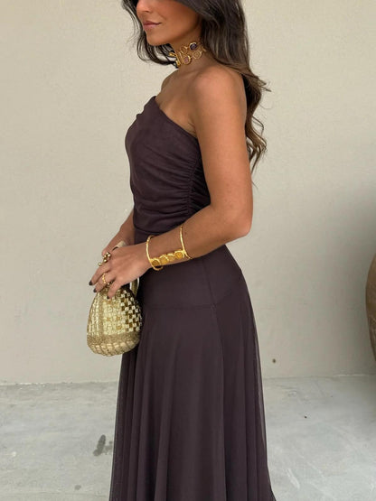 Brown - A woman wearing a dark brown, formal off-the-shoulder maxi dress featuring a ruched neckline and a flowing sheer mesh overlay over a fitted underlay, styled with gold accessories.