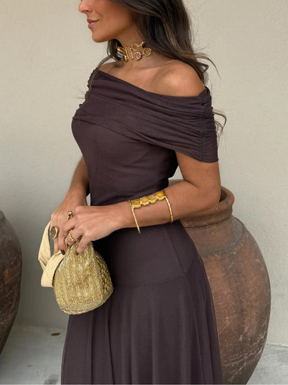 Brown - A woman wearing a dark brown, formal off-the-shoulder maxi dress featuring a ruched neckline and a flowing sheer mesh overlay over a fitted underlay, styled with gold accessories.