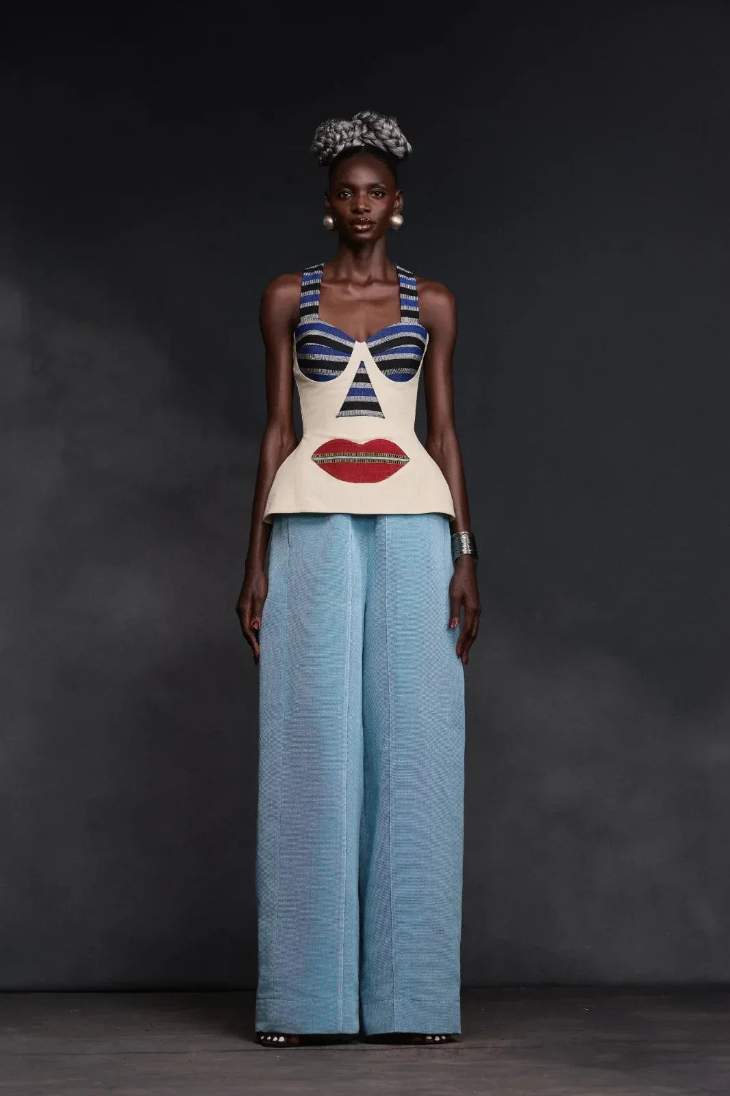 Multi - A woman wearing a structured, cream-colored sleeveless peplum top featuring an avant-garde surrealist face design with striped bust cups and a large red lip motif.