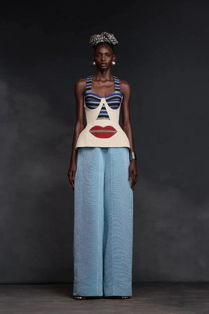 Multi - A woman wearing a structured, cream-colored sleeveless peplum top featuring an avant-garde surrealist face design with striped bust cups and a large red lip motif.