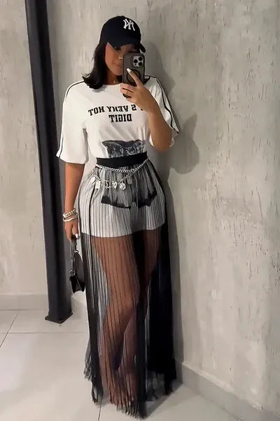 A woman taking a mirror selfie wearing a sheer black vertically pleated maxi skirt layered over light shorts, paired with a graphic t-shirt and chain belt.