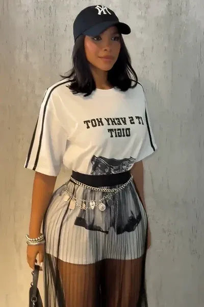 Top - A woman taking a mirror selfie wearing a sheer black vertically pleated maxi skirt layered over light shorts, paired with a graphic t-shirt and chain belt.