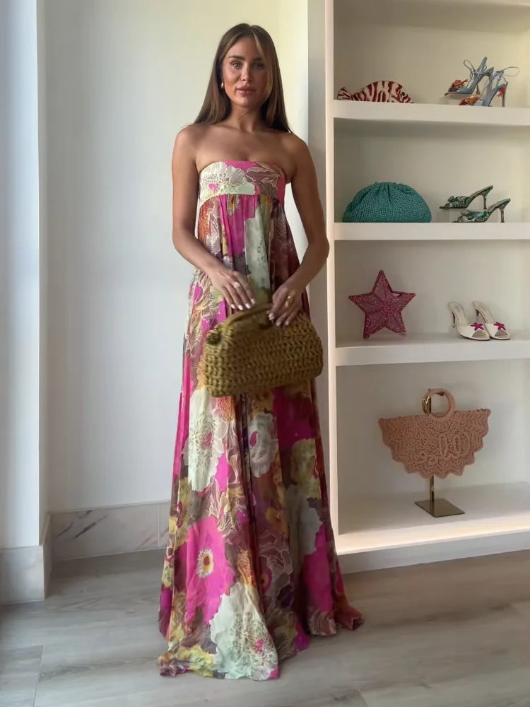 A woman wearing a strapless, flowing maxi dress featuring a vibrant, large-scale floral print in shades of pink, yellow, and green.