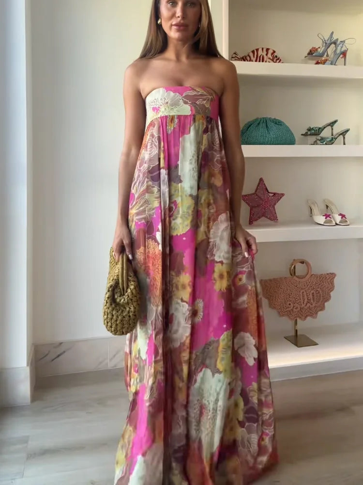 A woman wearing a strapless, flowing maxi dress featuring a vibrant, large-scale floral print in shades of pink, yellow, and green.