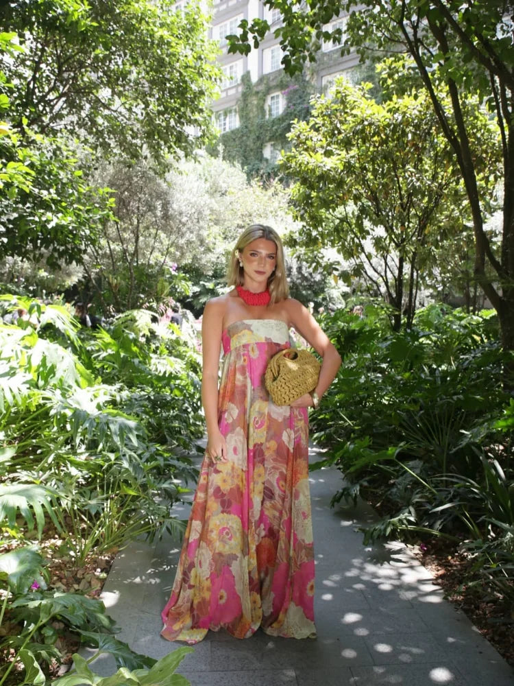 A woman wearing a strapless, flowing maxi dress featuring a vibrant, large-scale floral print in shades of pink, yellow, and green.