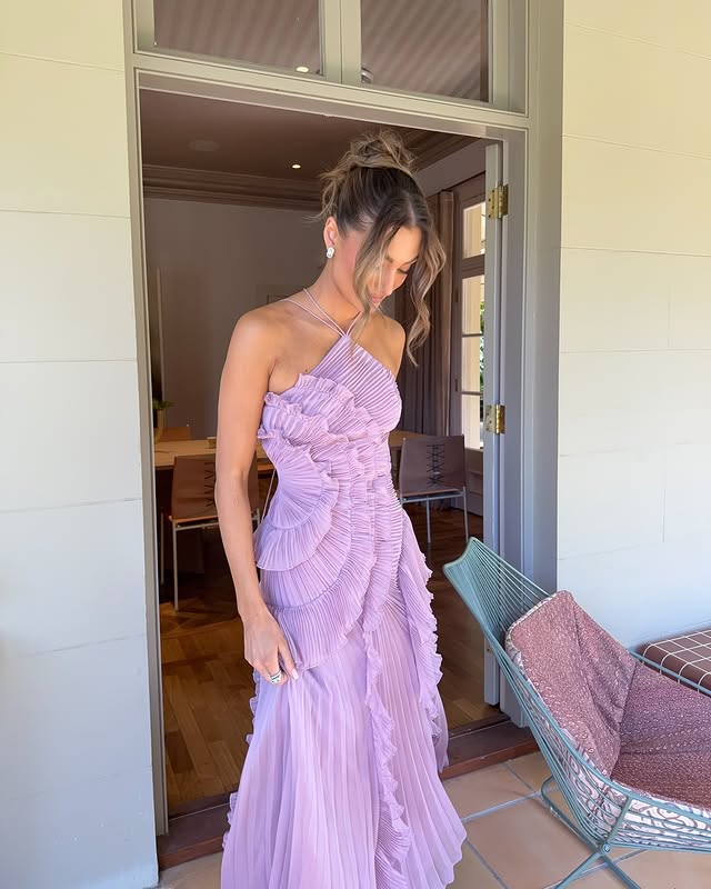 White - A woman wearing an elegant light purple midi dress featuring a halter neckline, all-over micro-pleating, and a dramatic asymmetrical cascading ruffle detail down the front.