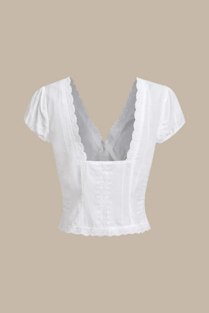 White - A woman wearing a white, short-sleeved corset-style blouse featuring a deep V-neckline with scalloped lace trim, a tie-front detail, and subtle tonal embroidery.