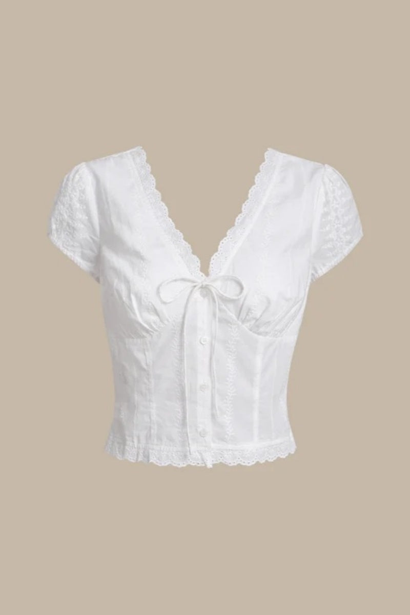 White - A woman wearing a white, short-sleeved corset-style blouse featuring a deep V-neckline with scalloped lace trim, a tie-front detail, and subtle tonal embroidery.