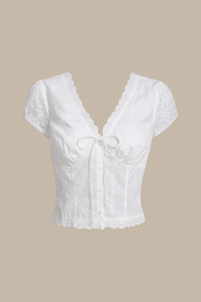 White - A woman wearing a white, short-sleeved corset-style blouse featuring a deep V-neckline with scalloped lace trim, a tie-front detail, and subtle tonal embroidery.