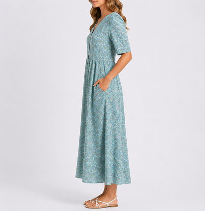 A woman wearing a woven floral smock midi dress with short sleeves and a gathered waist, standing with her hands in the side pockets against a plain light background.