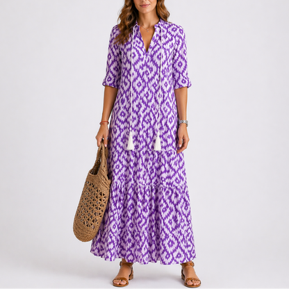 Purple - Woman wearing a woven ikat pattern maxi dress with a V-neck, tassel ties, 3/4-sleeves, and a tiered hemline against a light grey background.